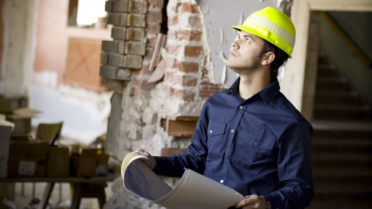 A construction worker or plumber in a yellow hard hat examines blueprints inside a partially demolished building.

He looks upward, appearing focused and thoughtful.