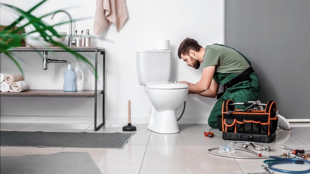 A florida plumber in green overalls kneels beside a toilet, concentrating on repairs. Nearby, tools are scattered on the floor, and a sink with toiletries is visible.
